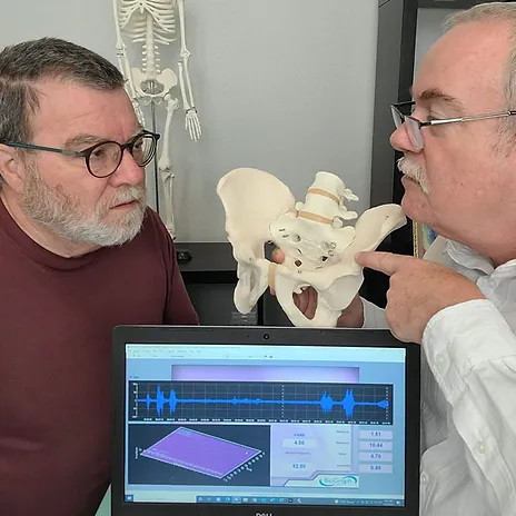 Two men examining a pelvic bone model; one is pointing at it while the other watches intently. A laptop displaying a chart and data is in the foreground. A skeleton model is in the background, representing Davenport Pelvic Therapy's commitment to detailed anatomical study.