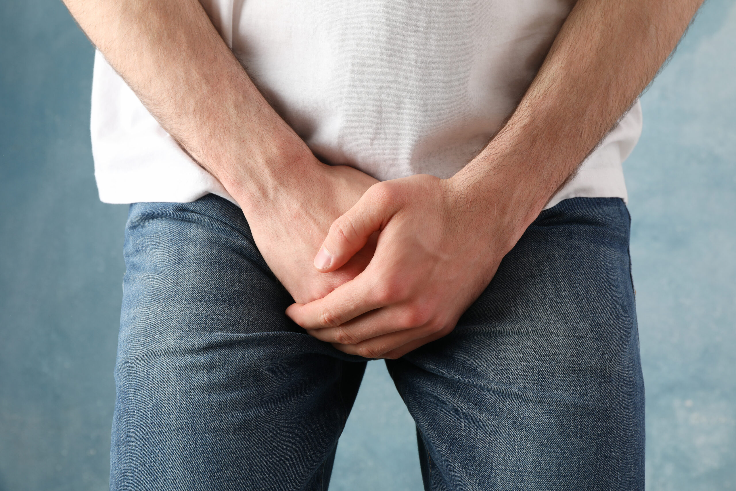 A person in a white shirt and blue jeans, perhaps considering incontinence treatment in Orlando, is standing with their hands clasped over their groin area against a blue background.