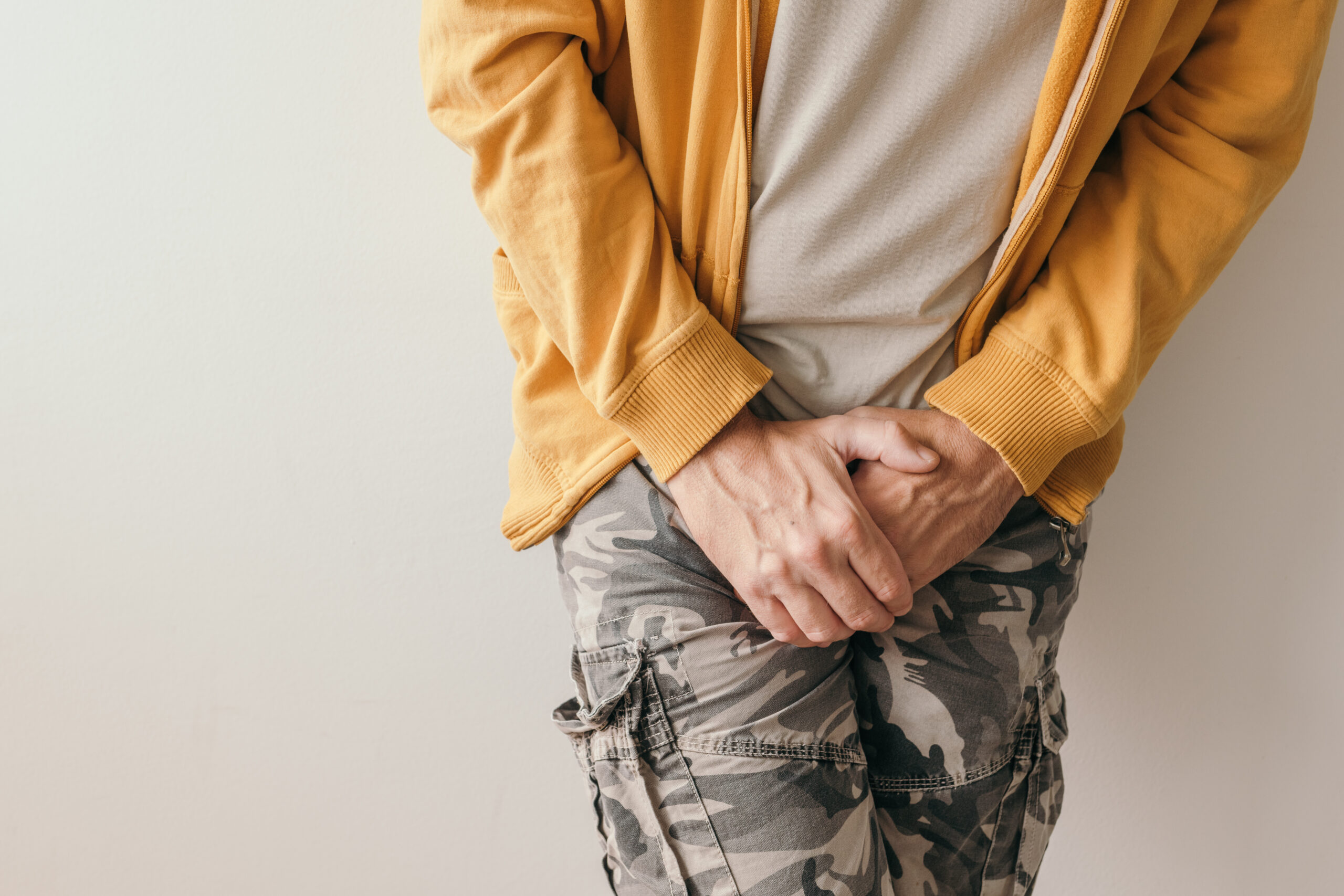 A person in a yellow jacket and camouflage pants holds their groin area while standing against a plain background, possibly needing incontinence treatment in Orlando, FL.