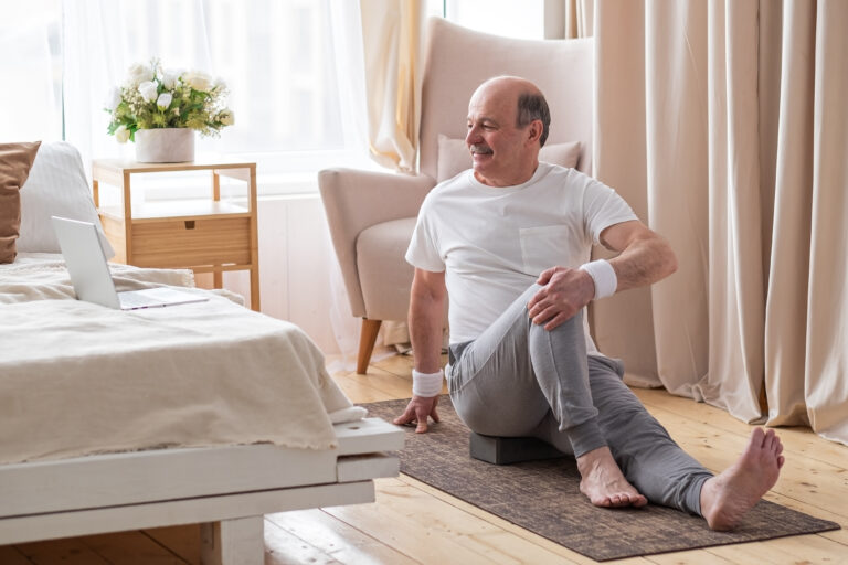 An older man sits on a yoga mat in a softly lit bedroom, stretching his legs while looking at a laptop. Dressed in comfortable workout clothes, he focuses on maintaining his health, perhaps with guidance from an FL Men's Health Clinic in Orlando. A bouquet of flowers adorns the bedside table.